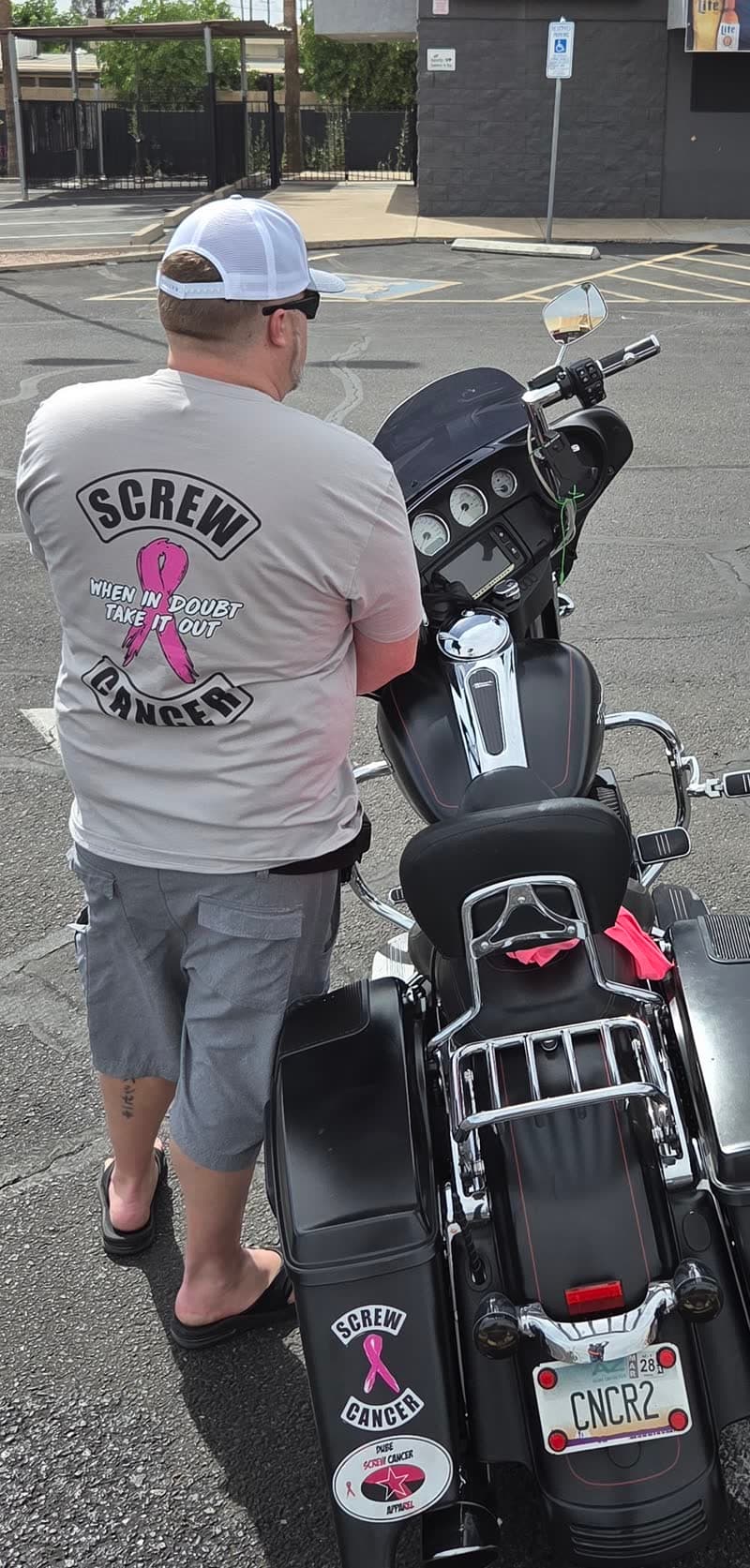 A community supporter wearing a pink-ribbon Screw Cancer shirt beside a motorcycle